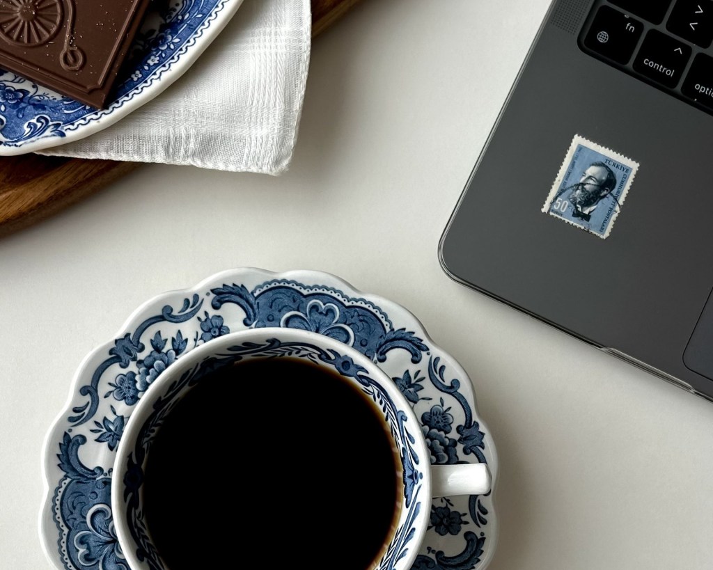 A cup of coffee, a computer and chocolates on a table.