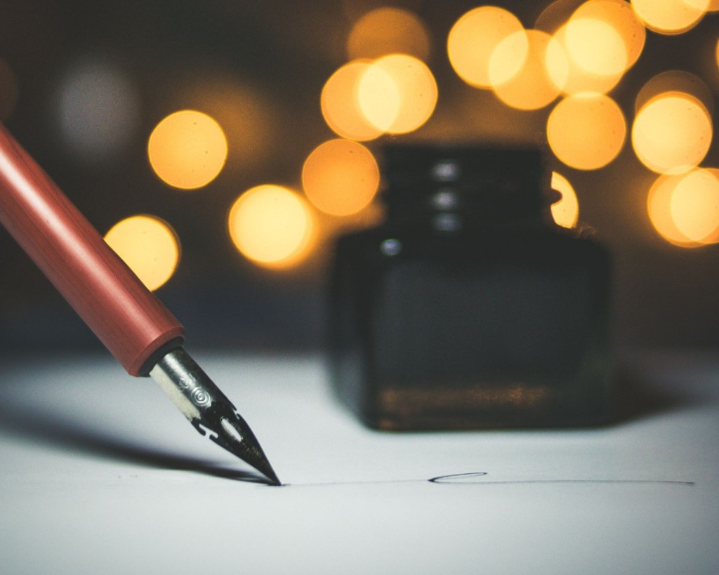 A dip pen writing on paper with an ink well and bokeh in the background.