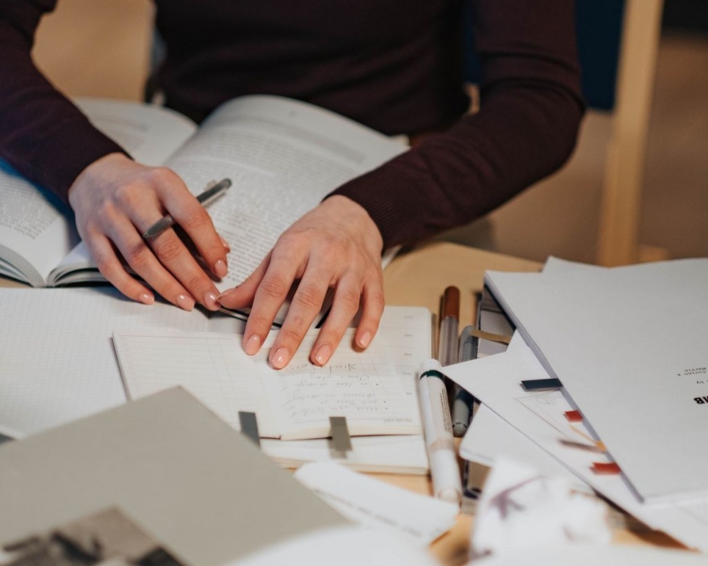A person holding a book open on a desk with a pen in hand among scattered papers.